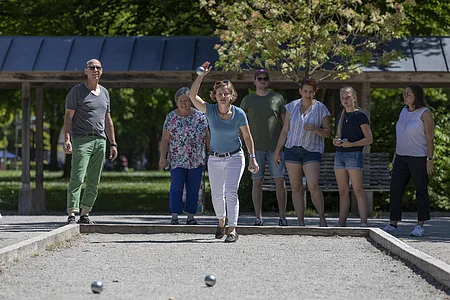 Sechs Personen spielen Boule auf einem Spielfeld im Freien, eine Frau wirft eine Kugel.