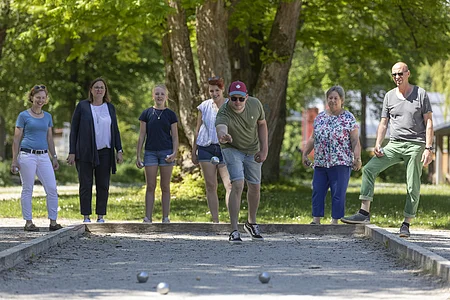 Sieben Personen spielen Boule auf einem Sandplatz im Park an einem sonnigen Tag.