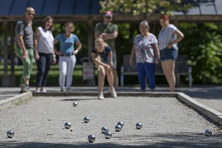 Gruppe von sieben Personen spielt Boule auf einem Sandplatz im Freien an einem sonnigen Tag.