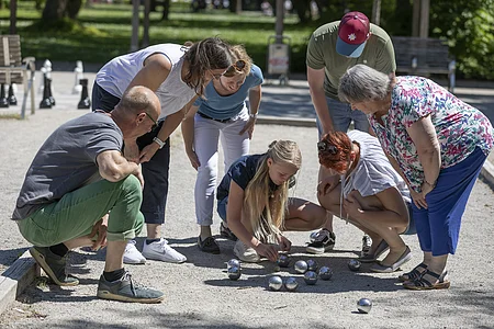 Sieben Personen bücken sich auf einem Boule-Spielfeld und betrachten die Metallkugeln auf dem Boden.