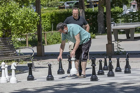 Zwei Männer spielen im Freien Schach mit übergroßen Figuren auf einem Schachbrett aus Steinplatten.