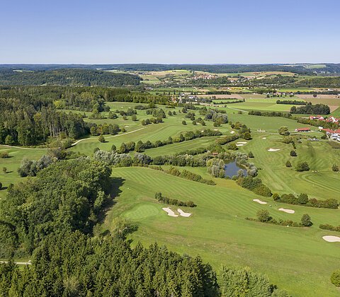 Luftaufnahme einer grünen Golfanlage mit Bäumen, Sandbunkern, Teich und Dorf im Hintergrund unter blauem Himmel.