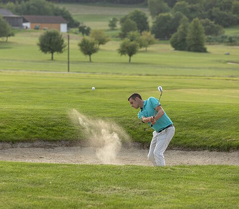 Mann spielt Golf und schlägt Ball aus einem Sandbunker auf einem Golfplatz mit Bäumen im Hintergrund.