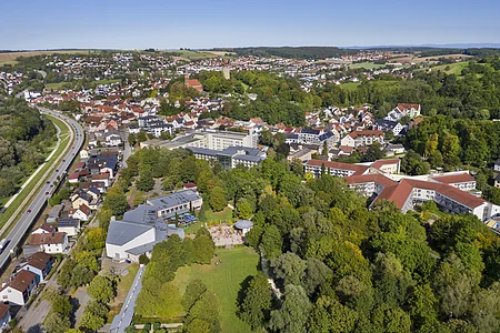 Luftaufnahme einer Stadt mit Wohnhäusern, Bäumen, einer Straße und Feldern im Hintergrund bei klarem Himmel.