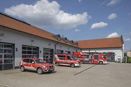 Feuerwehrfahrzeuge vor einem Feuerwehrhaus mit roten Dächern unter blauem Himmel mit Wolken.