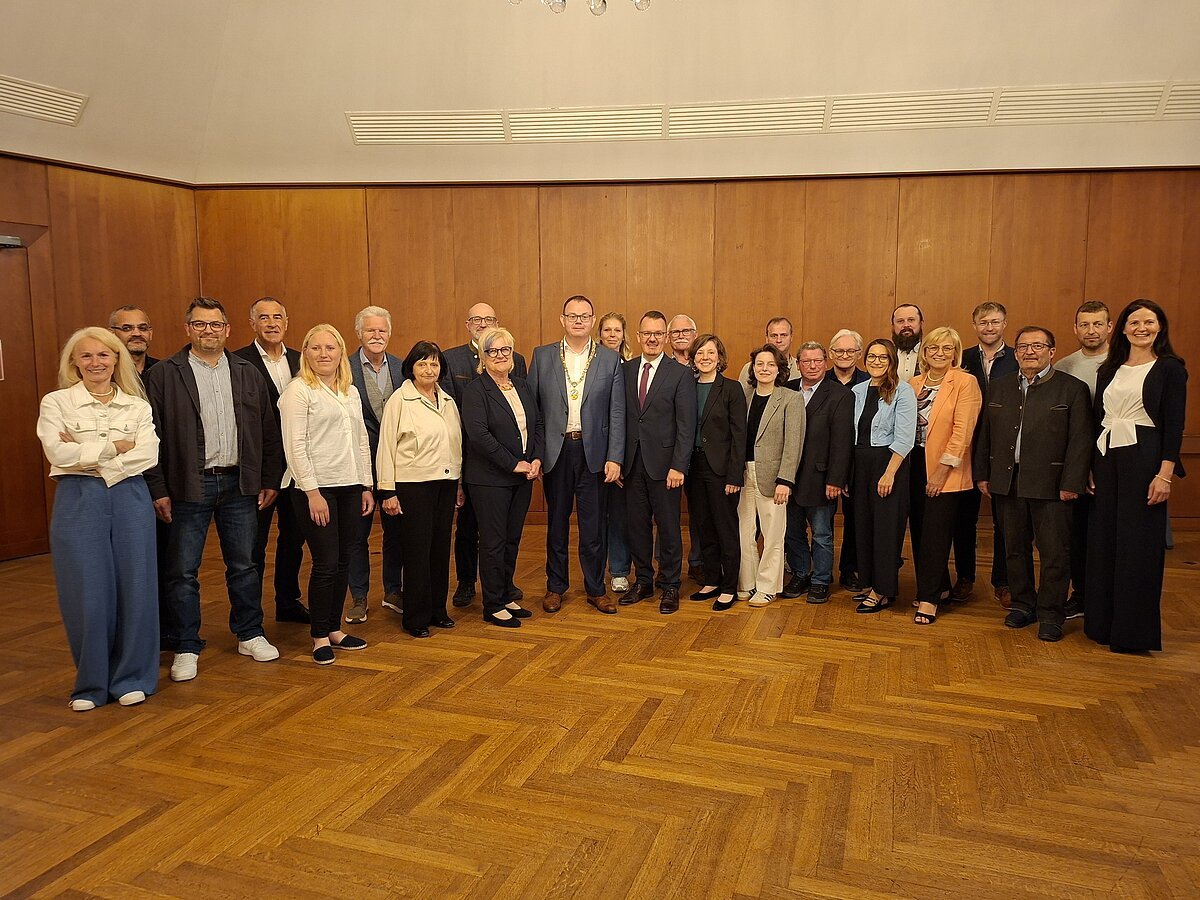Gruppenfoto von 26 Personen in einem Raum mit Holzwand und Parkettboden, alle stehen nebeneinander.