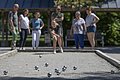 Sieben Personen spielen Boule auf einem Sandplatz, eine wirft gerade eine Kugel, im Vordergrund liegen mehrere Metallkugeln.