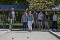 Sieben Personen spielen Boule auf einem Spielfeld im Freien bei Sonnenschein.