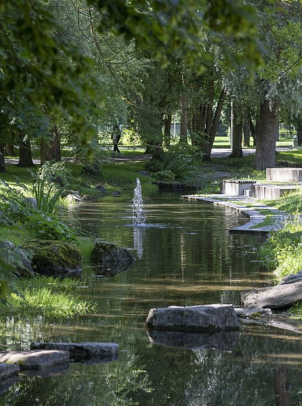 Blick auf einen schmalen Bach mit Springbrunnen in einem Park mit Bäumen, Wiesen und Sitzgelegenheiten.