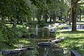 Blick auf einen schmalen Bach mit Springbrunnen in einem Park mit Bäumen, Wiesen und Sitzgelegenheiten.