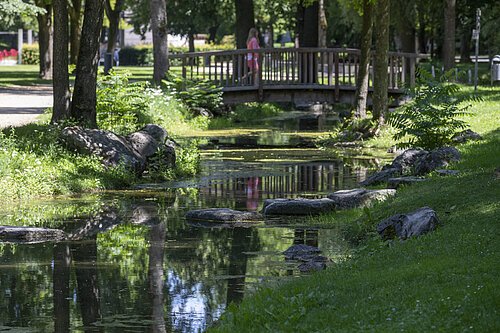 Markt Bad Abbach Kurpark Bachlauf Bach mit Steinen, Bäumen und Holzbrücke, Person in rosa Kleidung im Park im Hintergrund sichtbar.