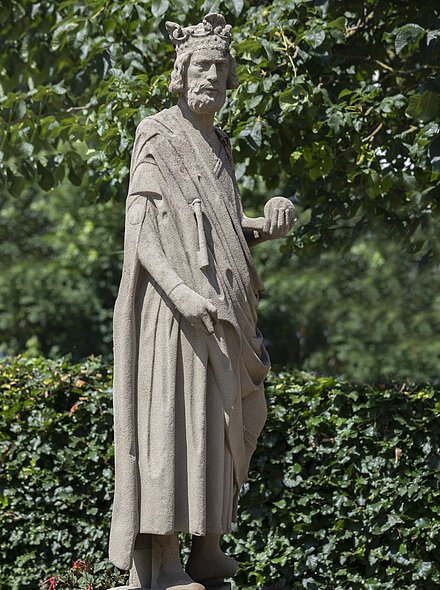 Markt Bad Abbach Figur Kaiser Heinrich II. Steinerne Statue eines bärtigen Königs mit Krone und Kugel in der rechten Hand, umgeben von grünen Pflanzen.