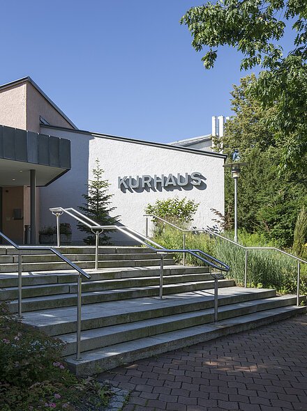 Haupteingang Kurhaus mit großem Veranstaltungssaal Eingang eines Gebäudes mit Treppe, Handläufen und dem Schriftzug „Kurhaus“ an der Fassade, umgeben von Bäumen und Sträuchern.
