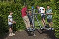 Fünf Jungen spielen an einem Outdoor-Fitnessgerät aus Metall in einem Park mit grünen Büschen.