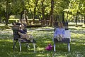 Zwei Frauen sitzen auf Liegestühlen auf einer Wiese mit Blumen, im Hintergrund Bäume und eine Holzbrücke.