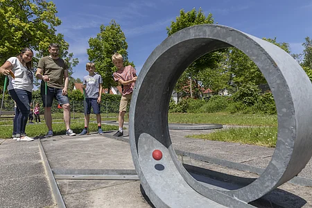Vier Personen spielen Minigolf auf einem sonnigen Außenplatz mit Betonhindernissen und grüner Umgebung.