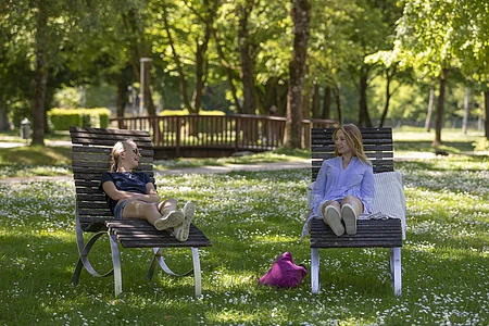 Zwei Frauen sitzen auf Liegestühlen auf einer Wiese mit Blumen, im Hintergrund Bäume und eine Holzbrücke.