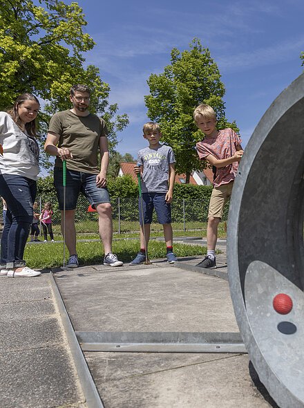 Vier Personen spielen Minigolf auf einem sonnigen Außenplatz mit Betonhindernissen und grüner Umgebung.