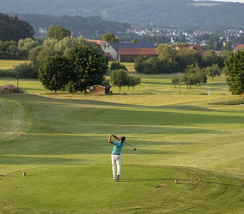 Golfer in türkisfarbenem Hemd schlägt Ball auf grünem Golfplatz mit Bäumen und Häusern im Hintergrund