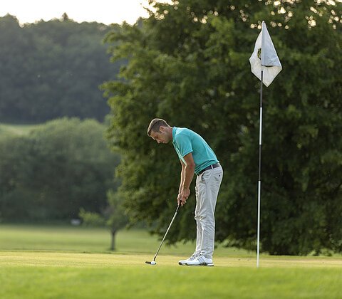 Mann in türkisfarbenem Hemd spielt Golf und steht neben Fahnenstock auf Golfplatz.