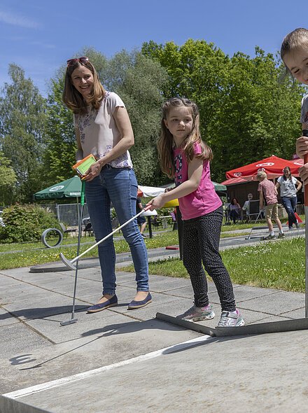 Drei Personen spielen Minigolf auf einem sonnigen Platz mit Bäumen und Sonnenschirmen im Hintergrund.