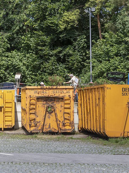 Vier gelbe Container mit der Aufschrift Demmel Sixtus AG stehen vor einem Wald, zwei Personen befüllen sie.