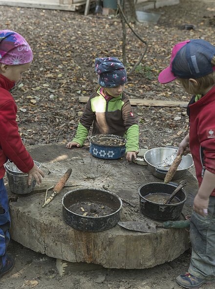 Drei Kinder spielen draußen an einem runden Holzstamm mit Töpfen und Kochutensilien im Wald.