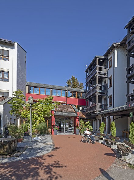 Außenansicht eines Seniorenwohnheims mit Sitzbänken, Rollstuhlfahrer und Personen auf Terrasse bei Sonnenschein.