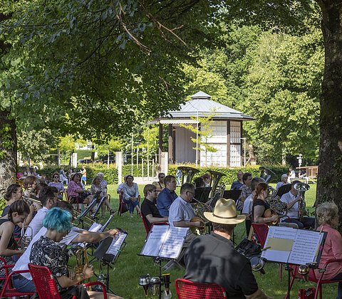 Bläserorchester spielt im Park auf roten Stühlen unter Bäumen mit Pavillon im Hintergrund.