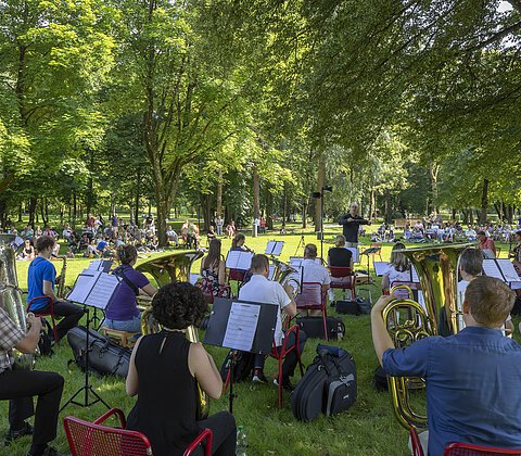 Blasorchester spielt im Park unter Bäumen, Publikum sitzt auf Wiese und Bänken im Hintergrund.