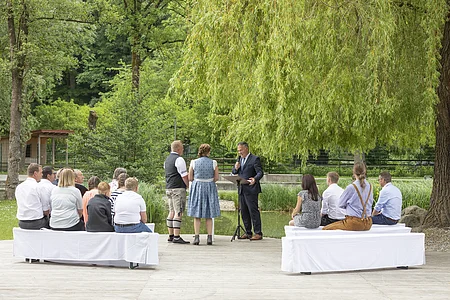Menschen sitzen auf weißen Bänken im Freien, ein Mann hält eine Rede vor einem Paar unter einem großen Baum.
