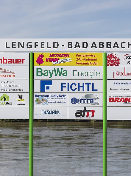 Werbetafel mit verschiedenen Firmenlogos und Hinweisen, Standort Lengfeld-Bad Abbach, Fluss im Hintergrund