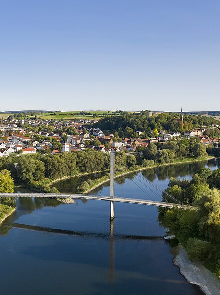 Fluss mit moderner Hängebrücke, umgeben von Bäumen und Häusern einer Kleinstadt unter blauem Himmel.