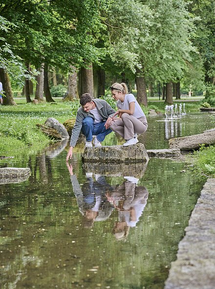 Zwei Jugendliche sitzen auf einem Stein inmitten des Baches im Kurpark inmitten der grünen Idylle.