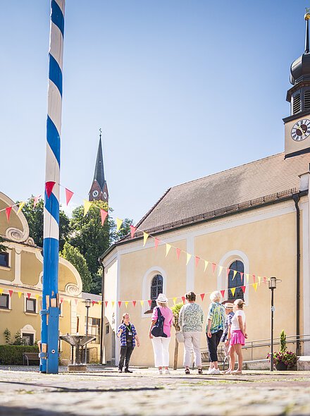 Vier erwachsene stehen in der Marktmitte neben der Marktkirche und folgen einem Gästeführer. Der Markt ist mit rot gelben Wimpeln dekoriert und links steht der blau-weiß gestreifte Maibaum steht unter blauem Himmel.