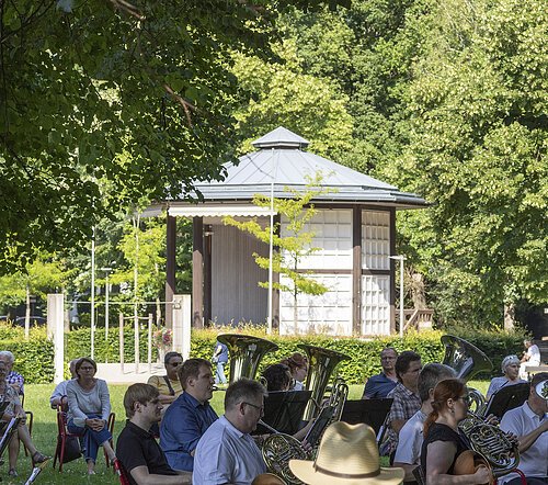 Bläserorchester spielt im Park auf roten Stühlen unter Bäumen mit Pavillon im Hintergrund.