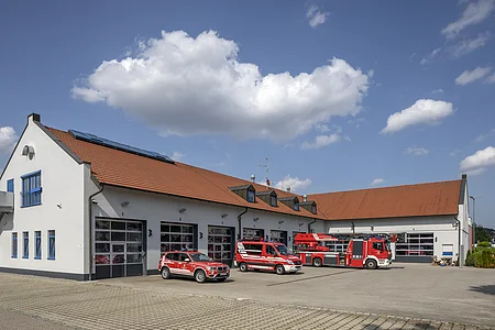 Feuerwache mit roten Einsatzfahrzeugen vor Garagentoren unter blauem Himmel mit Wolken.