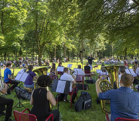 Orchester spielt im Park auf Wiese, Publikum sitzt auf Stühlen und Decken unter Bäumen.
