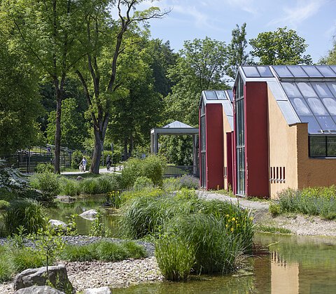 Park mit kleinem Teich, Büschen, Bäumen und zwei gelb-roten Gebäuden mit Glasdächern bei Sonnenschein