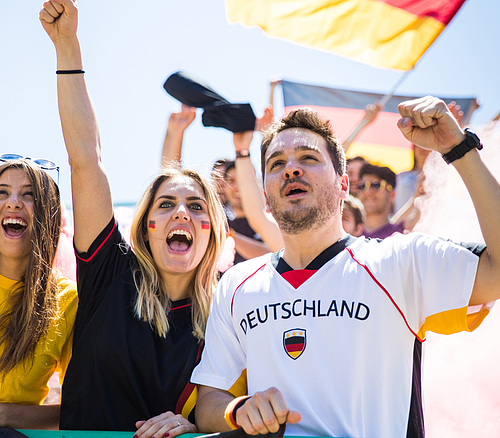 Jubelnde Fans mit Deutschland-Fahnen und Deutschland-Schriftzug auf Trikot bei Sportveranstaltung im Freien.