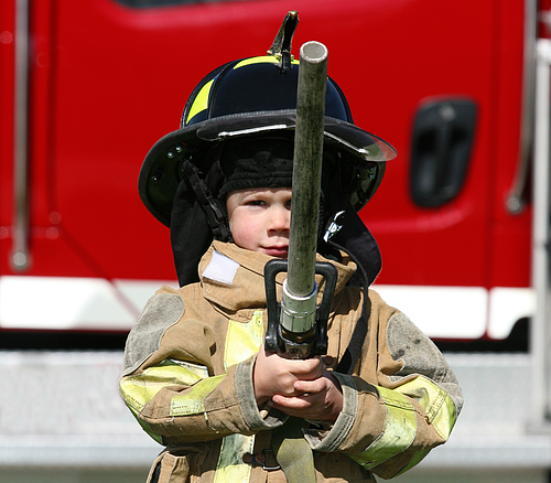 Kinderfeuerwehrmann Kind in Feuerwehrschutzkleidung hält Feuerwehrschlauch vor rotem Feuerwehrauto in Nahaufnahme