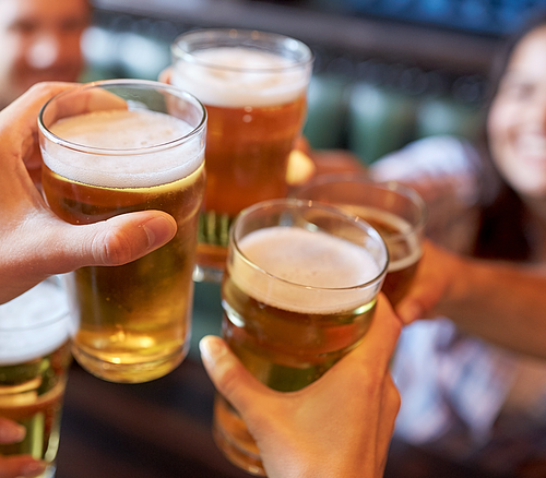 Freunde stoßen an. Mehrere Hände stoßen mit Biergläsern an, unscharfe Personen im Hintergrund.