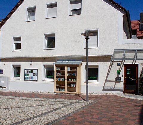 Weißes Gebäude mit Bücherschrank und mehreren Fenstern an einem gepflasterten Platz bei Sonnenschein