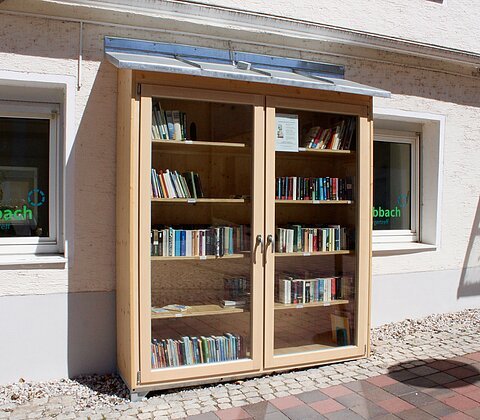 Holzschrank mit Glastüren und Büchern steht vor einem hellen Haus mit zwei Fenstern und Pflastersteinen.