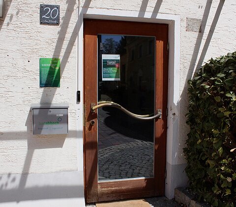 Eingangstür mit Glas und geschwungenem Griff, Hausnummer 20, Briefkasten und Busch rechts daneben.