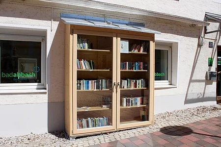 Holzschrank mit Glastüren und Büchern steht vor einem hellen Haus mit zwei Fenstern und Pflastersteinen.
