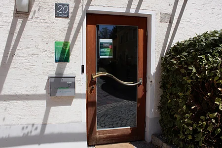 Eingangstür mit Glas und geschwungenem Griff, Hausnummer 20, Briefkasten und Busch rechts daneben.