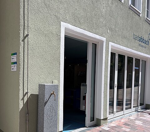 Eingang eines Gebäudes mit geöffnetem Glastür, grauer Wasserzapfstelle und gepflastertem Boden.