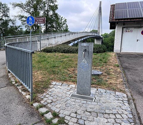 Trinkwasserbrunnen auf gepflastertem Boden neben einem Fuß- und Radweg bei der Fußgängerbrücke