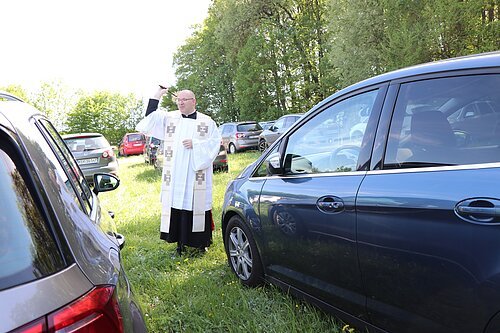 Priester in weißem Gewand segnet parkende Autos auf einer Wiese mit Bäumen im Hintergrund.